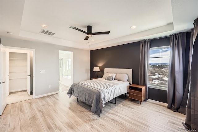 bedroom with a tray ceiling, ensuite bath, ceiling fan, and light wood-type flooring