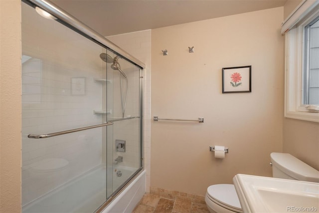 bathroom with stone finish floor, baseboards, toilet, and bath / shower combo with glass door