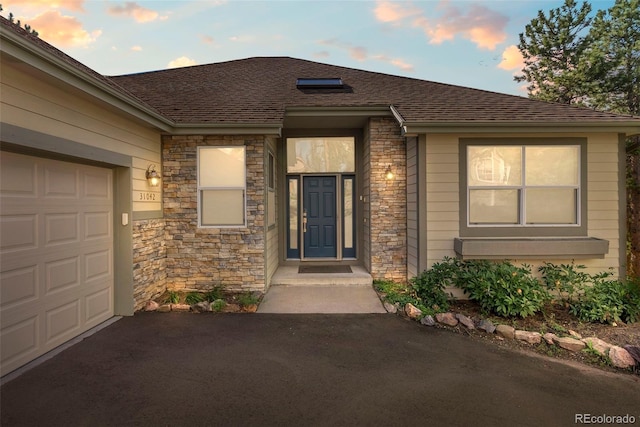 exterior entry at dusk featuring stone siding, a shingled roof, an attached garage, and driveway