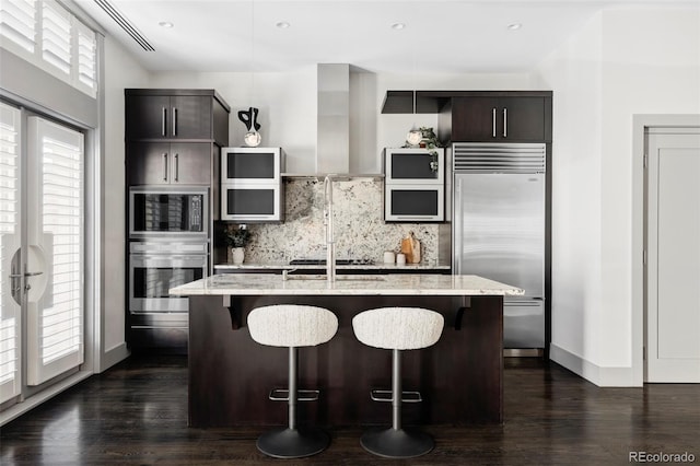 kitchen with light stone countertops, built in appliances, tasteful backsplash, a center island with sink, and a breakfast bar area