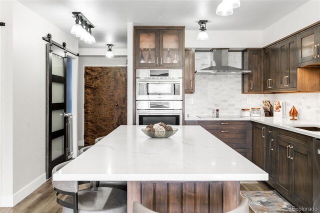 1551 Larimer St Unit 1405, Denver, CO 80202 - kitchen featuring decorative backsplash, dark brown cabinets, appliances with stainless steel finishes, a barn door, and wall chimney range hood
