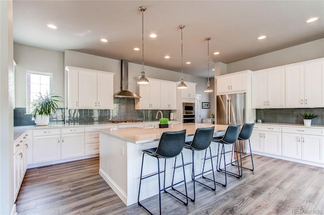 9571 Yucca Ct, Arvada, CO 80007 - kitchen with wall chimney range hood, decorative backsplash, an island with sink, stainless steel appliances, and light wood-style flooring