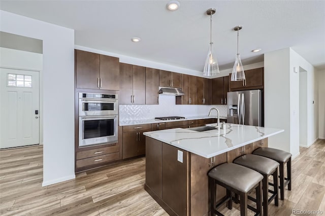 kitchen with a center island with sink, hanging light fixtures, stainless steel appliances, dark brown cabinets, and a sink