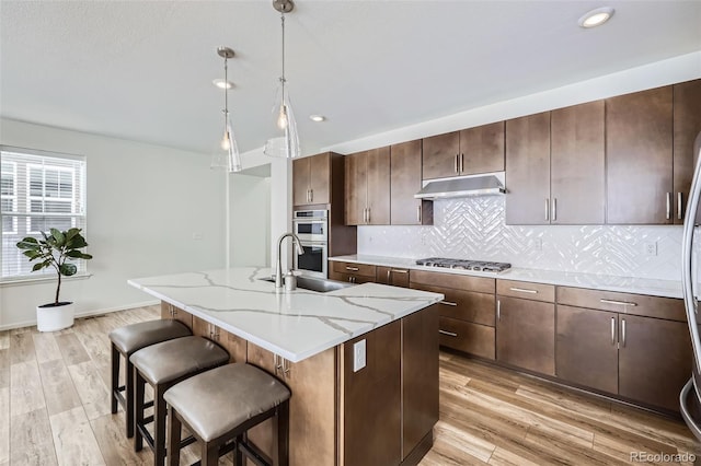 kitchen featuring stainless steel appliances, hanging light fixtures, a kitchen island with sink, a sink, and under cabinet range hood