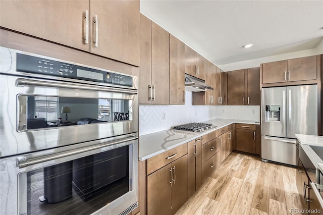 kitchen with backsplash, appliances with stainless steel finishes, light stone countertops, light wood-type flooring, and under cabinet range hood