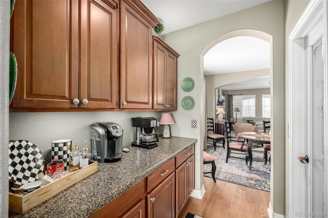 kitchen with brown cabinetry, light wood-type flooring, dark stone counters, and arched walkways