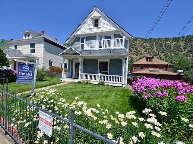 victorian house with a balcony, a porch, a front yard, and fence