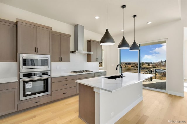 kitchen with sink, stainless steel appliances, wall chimney range hood, a center island with sink, and light wood-type flooring