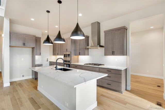 kitchen with light stone countertops, wall chimney range hood, an island with sink, appliances with stainless steel finishes, and light wood-type flooring