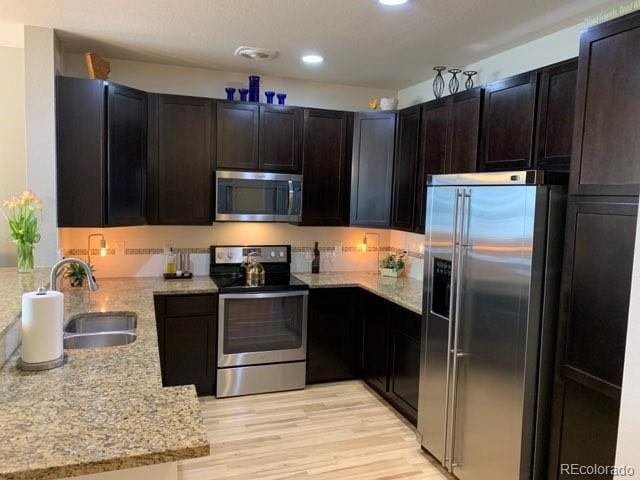 kitchen with sink, dark brown cabinets, light wood-type flooring, appliances with stainless steel finishes, and light stone countertops