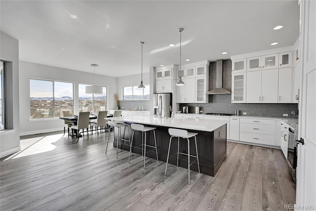 kitchen featuring light hardwood / wood-style flooring, white cabinets, wall chimney range hood, a kitchen island with sink, and stainless steel refrigerator with ice dispenser