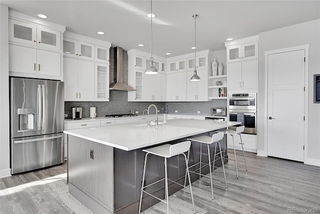 kitchen featuring tasteful backsplash, stainless steel appliances, a center island with sink, white cabinetry, and wall chimney exhaust hood