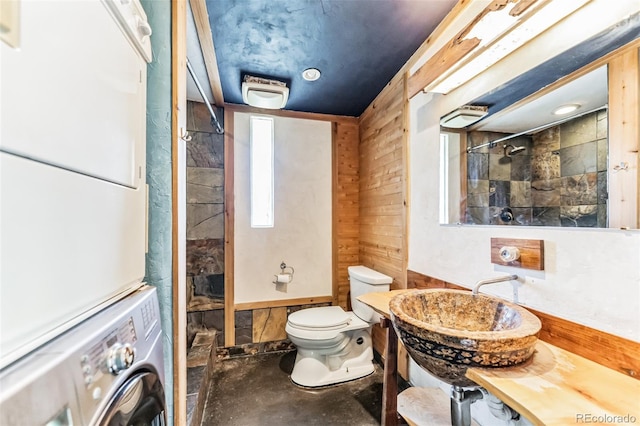 bathroom featuring sink, wooden walls, toilet, stacked washing maching and dryer, and tiled shower