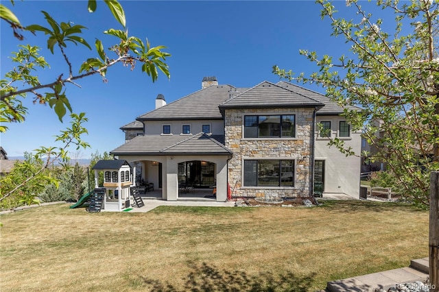 rear view of house featuring stone siding, a chimney, a patio area, a playground, and a lawn