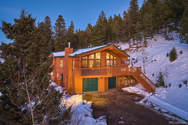 snow covered property featuring a garage, dirt driveway, a view of trees, a chimney, and stairway