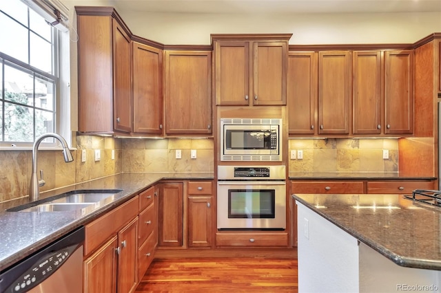 kitchen with appliances with stainless steel finishes, a sink, light wood finished floors, dark stone countertops, and decorative backsplash
