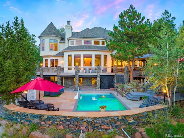 back of property at dusk with stairs, a chimney, a patio area, stone siding, and an outdoor pool