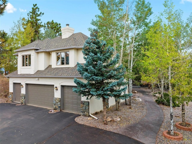 view of front facade featuring an attached garage, a shingled roof, a chimney, stucco siding, and aphalt driveway