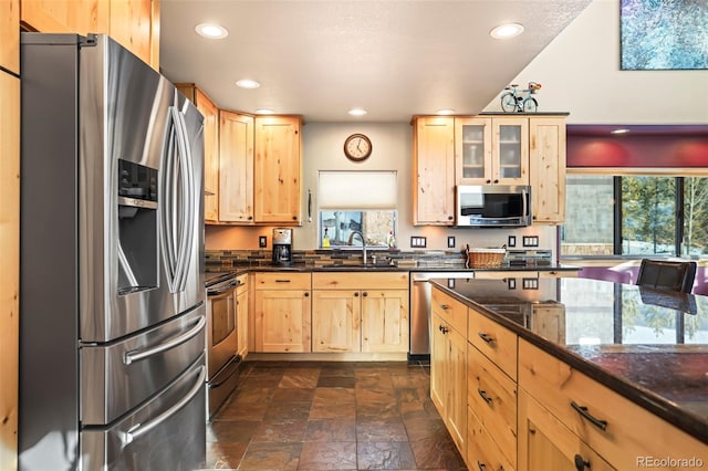 kitchen featuring dark stone countertops, light brown cabinets, a sink, stainless steel appliances, and glass insert cabinets