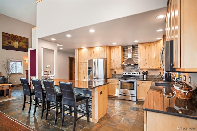 kitchen featuring light brown cabinets, a breakfast bar, a sink, stainless steel appliances, and wall chimney range hood