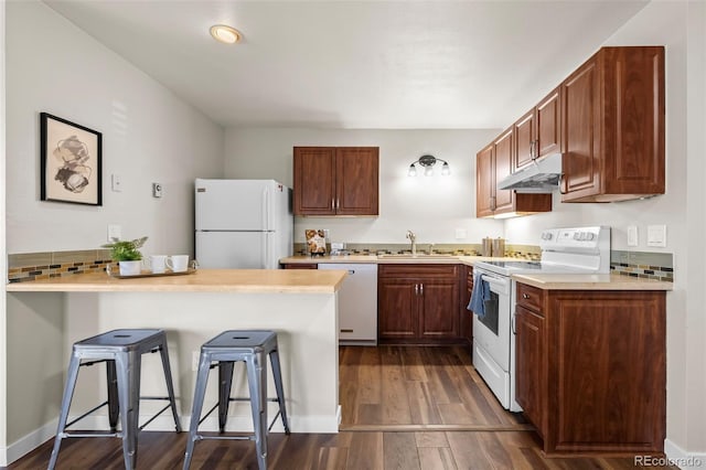 kitchen featuring a sink, a breakfast bar, under cabinet range hood, white appliances, and dark wood-style flooring
