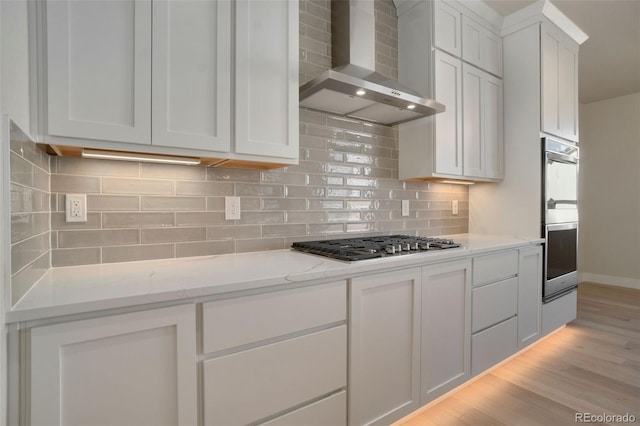 kitchen with wall chimney range hood, appliances with stainless steel finishes, light stone countertops, white cabinets, and light wood-type flooring