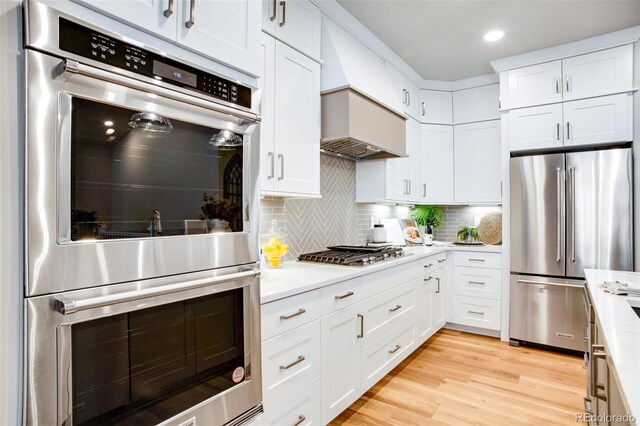 kitchen with white cabinetry, decorative backsplash, appliances with stainless steel finishes, custom range hood, and light hardwood / wood-style floors