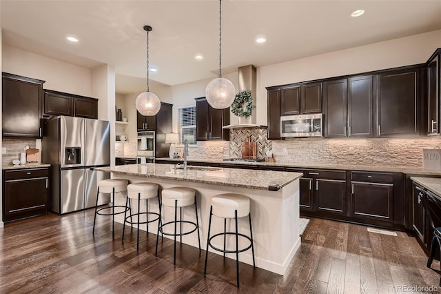 kitchen featuring appliances with stainless steel finishes, wall chimney exhaust hood, decorative light fixtures, dark hardwood / wood-style floors, and an island with sink