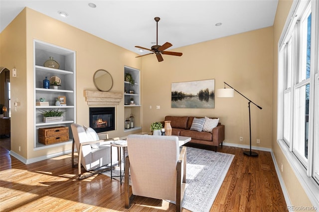 living area with ceiling fan, baseboards, built in features, wood finished floors, and a glass covered fireplace