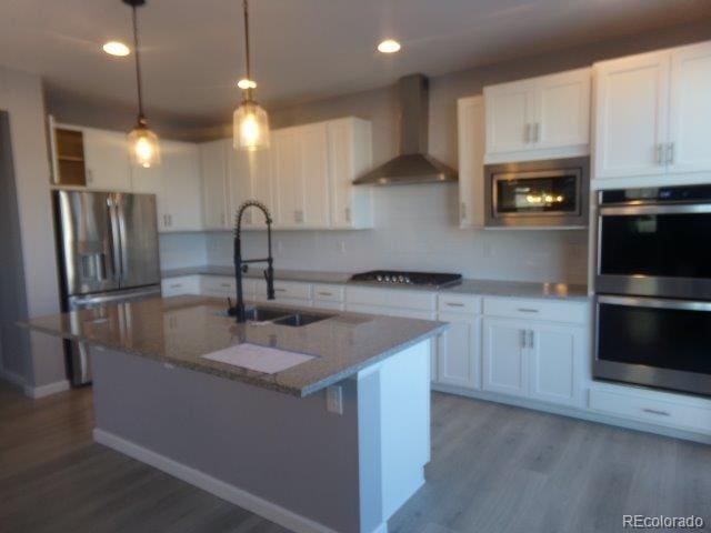 kitchen with sink, appliances with stainless steel finishes, wall chimney range hood, a kitchen island with sink, and white cabinets