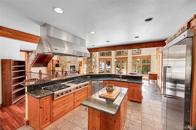 kitchen with appliances with stainless steel finishes, a peninsula, a stone fireplace, wall chimney range hood, and a sink