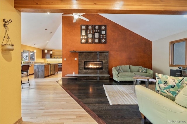 living area featuring ceiling fan, high vaulted ceiling, a stone fireplace, beam ceiling, and light wood finished floors