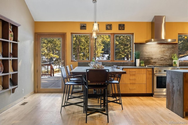 kitchen with light wood finished floors, decorative backsplash, high end stove, wall chimney range hood, and pendant lighting