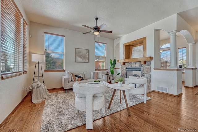 living area with visible vents, ceiling fan, a stone fireplace, wood finished floors, and ornate columns