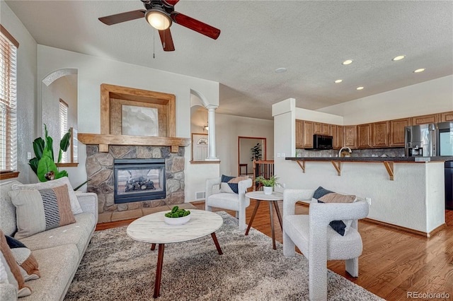 living room featuring ceiling fan, a stone fireplace, light wood-style flooring, a textured ceiling, and ornate columns