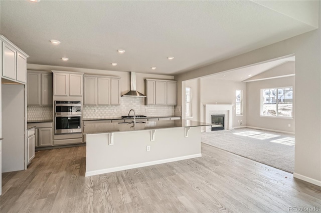 kitchen featuring gray cabinetry, lofted ceiling, a kitchen island with sink, wall chimney range hood, and a breakfast bar area