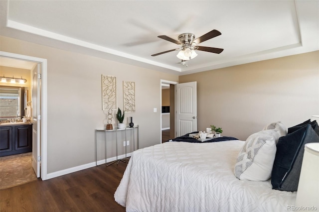 bedroom with dark wood-style floors, a tray ceiling, ensuite bath, baseboards, and ceiling fan