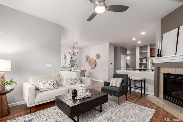 living area featuring a ceiling fan, wood finished floors, baseboards, a tiled fireplace, and a chandelier