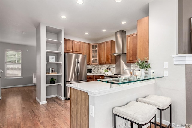 kitchen featuring freestanding refrigerator, wall chimney exhaust hood, brown cabinetry, a peninsula, and open shelves
