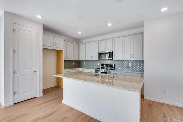 kitchen featuring stainless steel appliances, an island with sink, backsplash, light stone counters, and baseboards