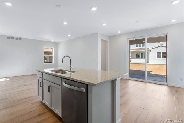 kitchen with stainless steel dishwasher, a sink, light wood-style floors, light stone countertops, and a center island with sink
