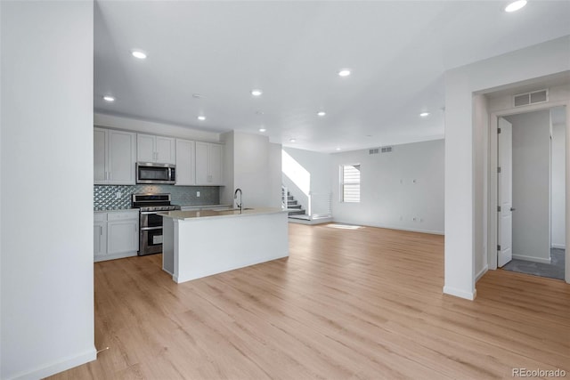 kitchen featuring stainless steel appliances, sink, backsplash, a kitchen island with sink, and light wood-type flooring