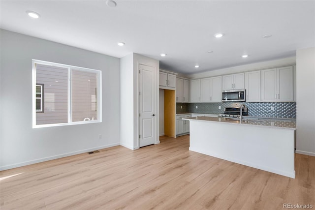 kitchen with stainless steel microwave, light wood finished floors, light countertops, a kitchen island with sink, and recessed lighting