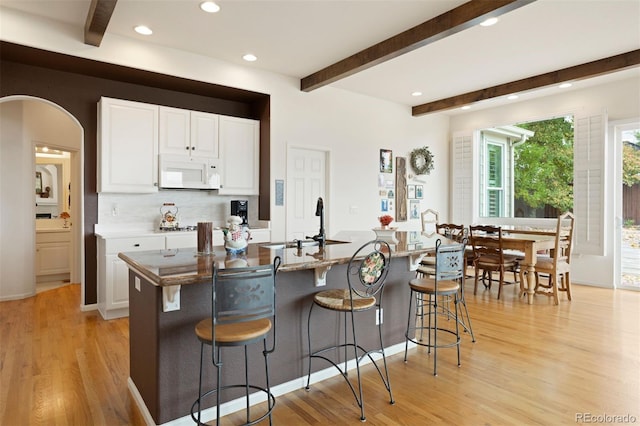 kitchen with beam ceiling, a kitchen island with sink, white cabinets, and light wood-type flooring