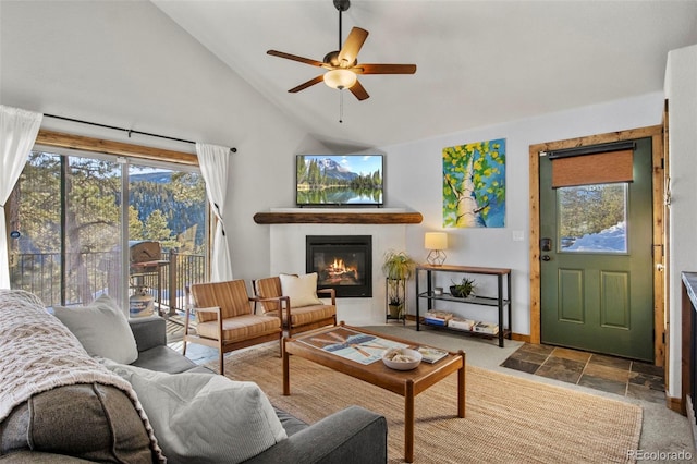 living room featuring stone finish flooring, high vaulted ceiling, a glass covered fireplace, and a ceiling fan