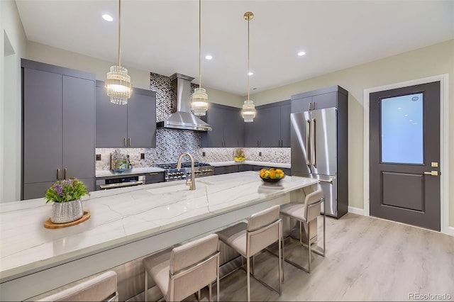 kitchen with light stone counters, wall chimney range hood, high end refrigerator, light wood-type flooring, and pendant lighting