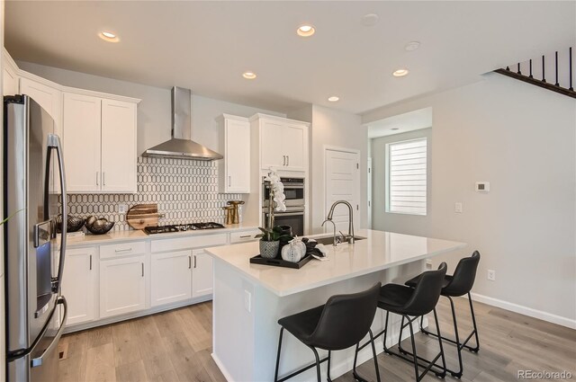 kitchen with wall chimney range hood, white cabinets, a kitchen island with sink, light wood-type flooring, and stainless steel appliances