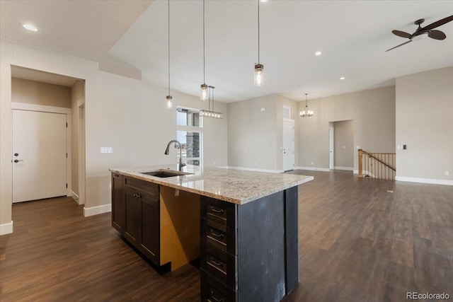 kitchen with sink, hanging light fixtures, a kitchen island with sink, light stone counters, and dark wood-type flooring