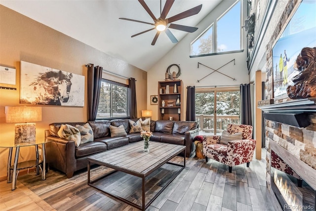 living area featuring high vaulted ceiling, ceiling fan, a stone fireplace, and wood finished floors