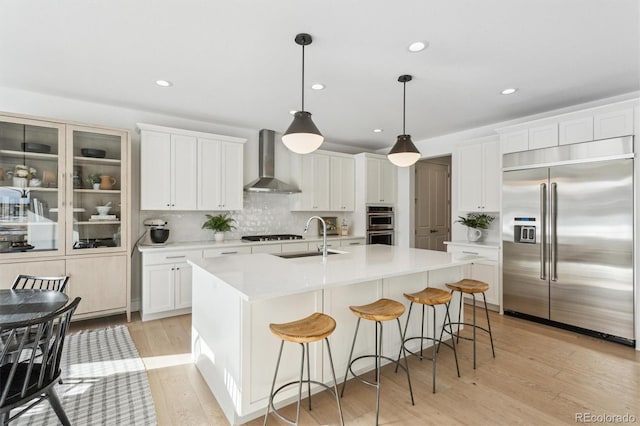 kitchen featuring wall chimney range hood, sink, appliances with stainless steel finishes, white cabinetry, and a center island with sink